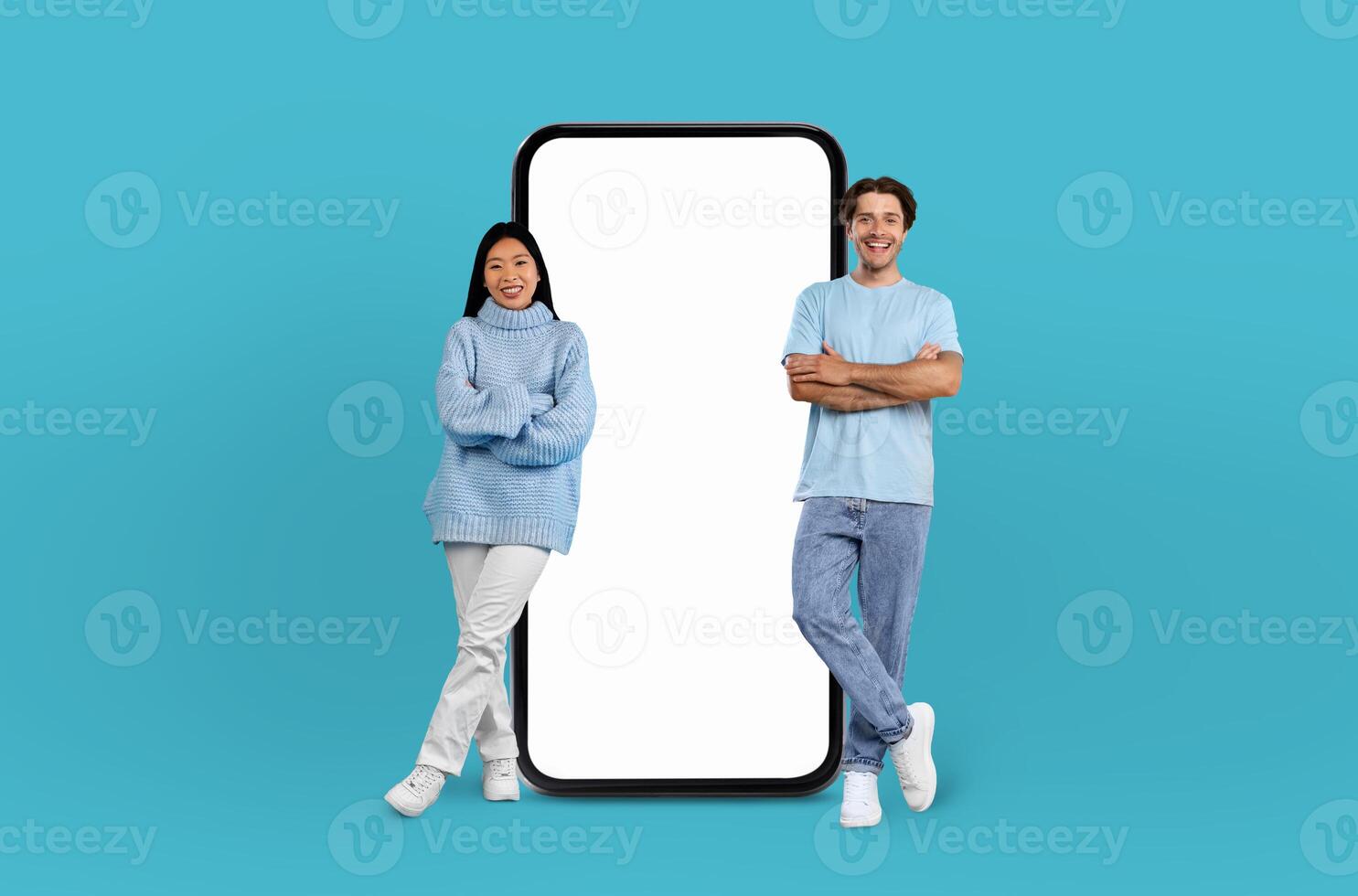 On a deep blue background, a multiethnic couple, both smiling, stands adjacent to an enormous smartphone with a blank display, ready for showcasing apps or designs. photo