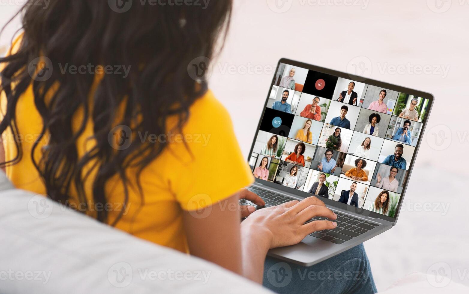 Black woman with long, black hair sits on a couch while participating in an online meeting, using a laptop with a screen showing a grid of multiple feeds of other participants in the meeting. photo