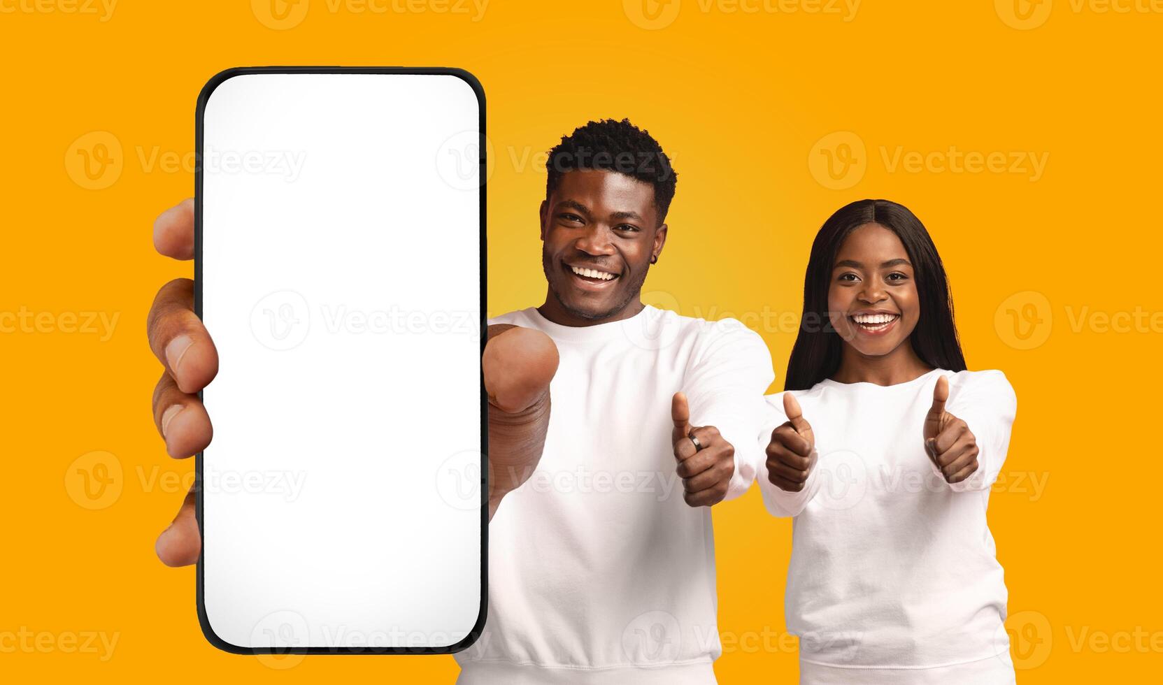A young man and woman, both African American and smiling, stand side-by-side in front of a bright yellow background. The man holds a smartphone with a blank white screen photo