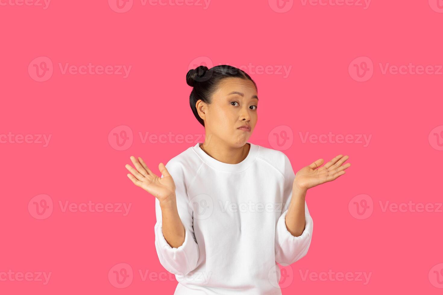 Asian young woman in casual attire raises her hands in confusion, expressing uncertainty with a lighthearted posture and a soft smile. photo