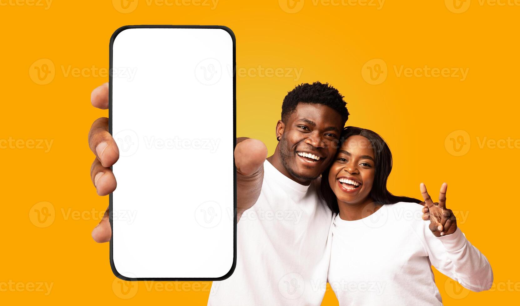 A young black couple smiles brightly as they hold a smartphone with a blank white screen in front of them. The woman makes a peace sign with her hand, while the man points at the screen. photo