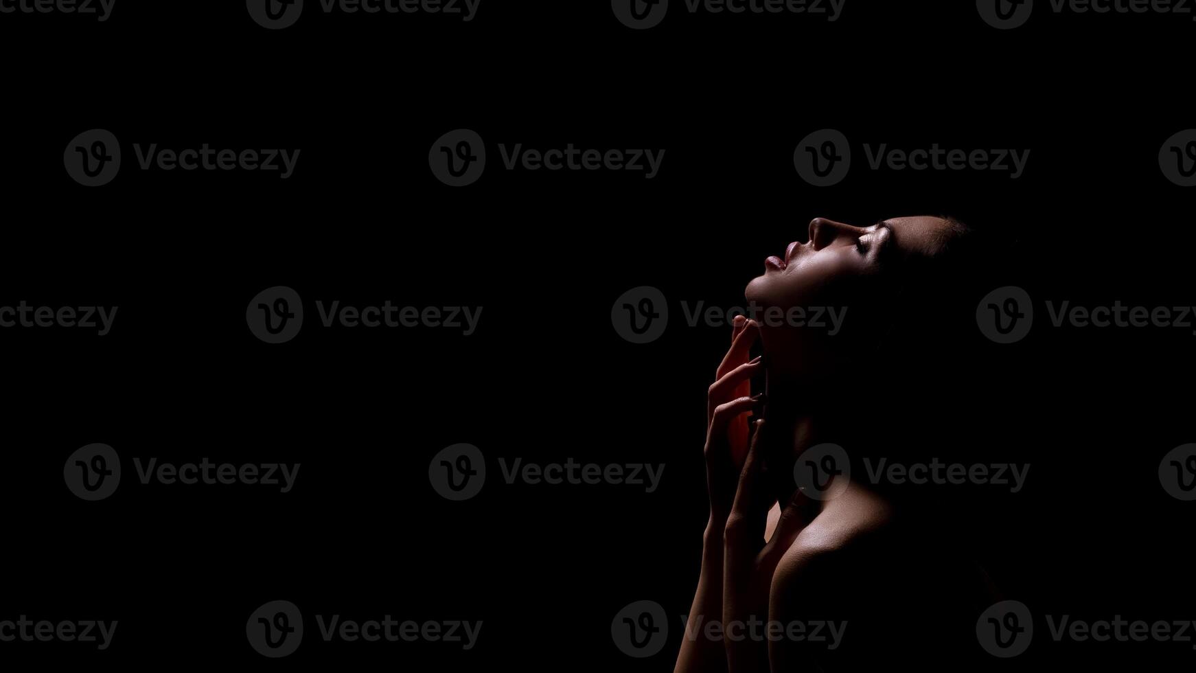 A woman with her head tilted back, looking towards the top of the frame, is silhouetted against a dark background. She has her fingers gently on her neck, copy space photo