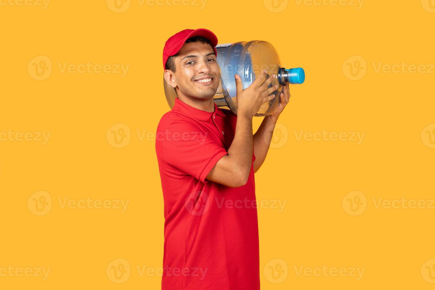 A cheerful young delivery man in a red uniform and cap holds a large water container on his shoulder. The bright yellow background adds a vibrant feel to the scene photo