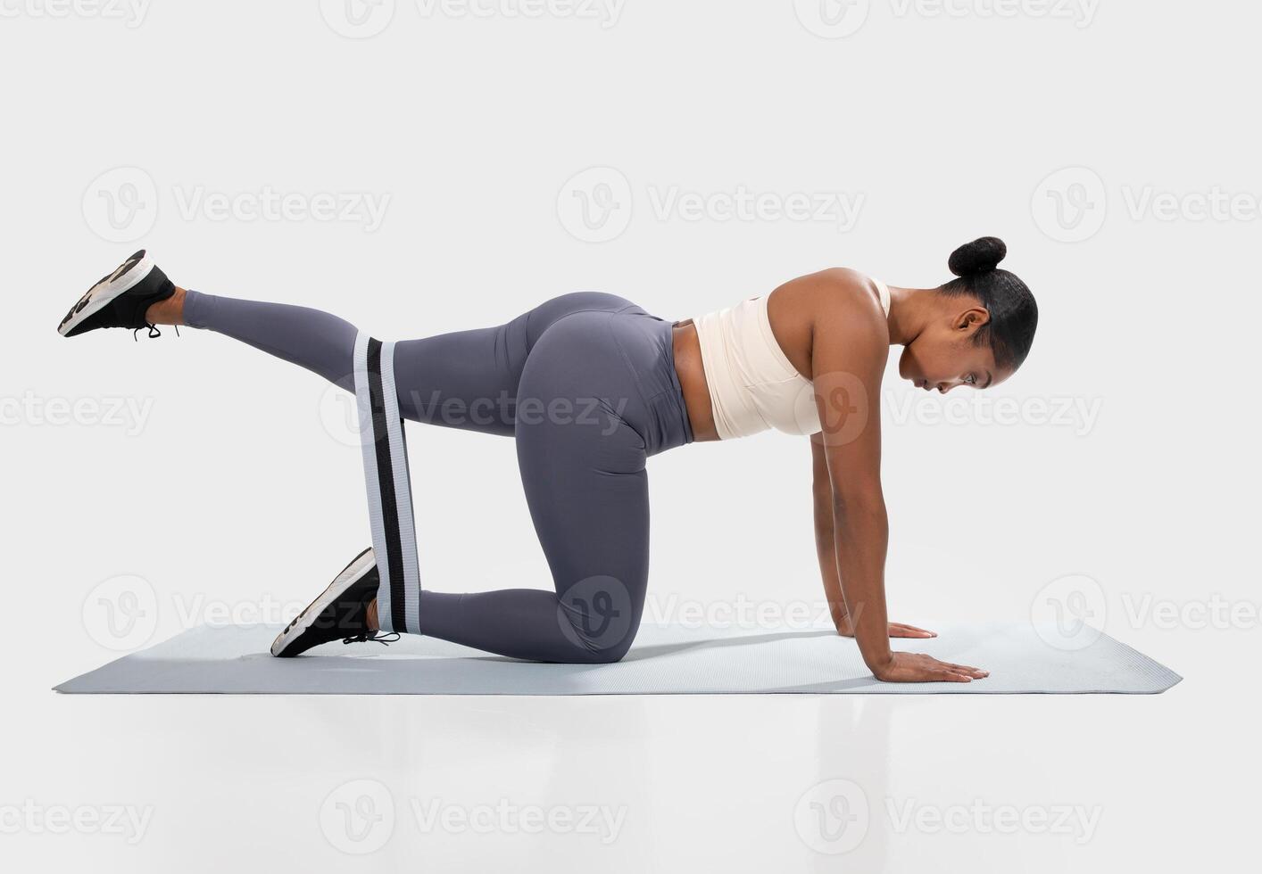 African American woman in a white sports bra and grey leggings is performing a glute bridge exercise on a blue yoga mat. She is using a black and white resistance band around her upper thigh photo