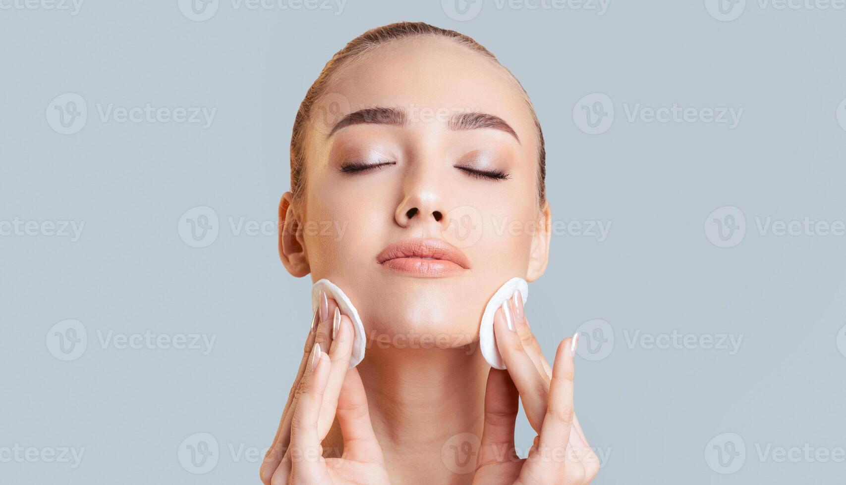 A woman with her eyes closed gently applies cotton pads to her face as part of her skincare routine. She is smiling softly, and her skin is clear and healthy. photo