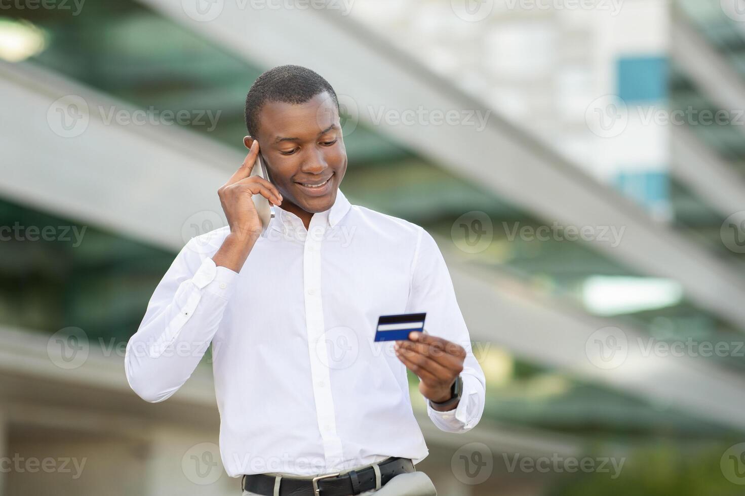 African American man stands outdoors, smiling as he examines his credit card while conversing on his mobile phone. He is engaged, possibly discussing financial matters or making a purchase decision photo