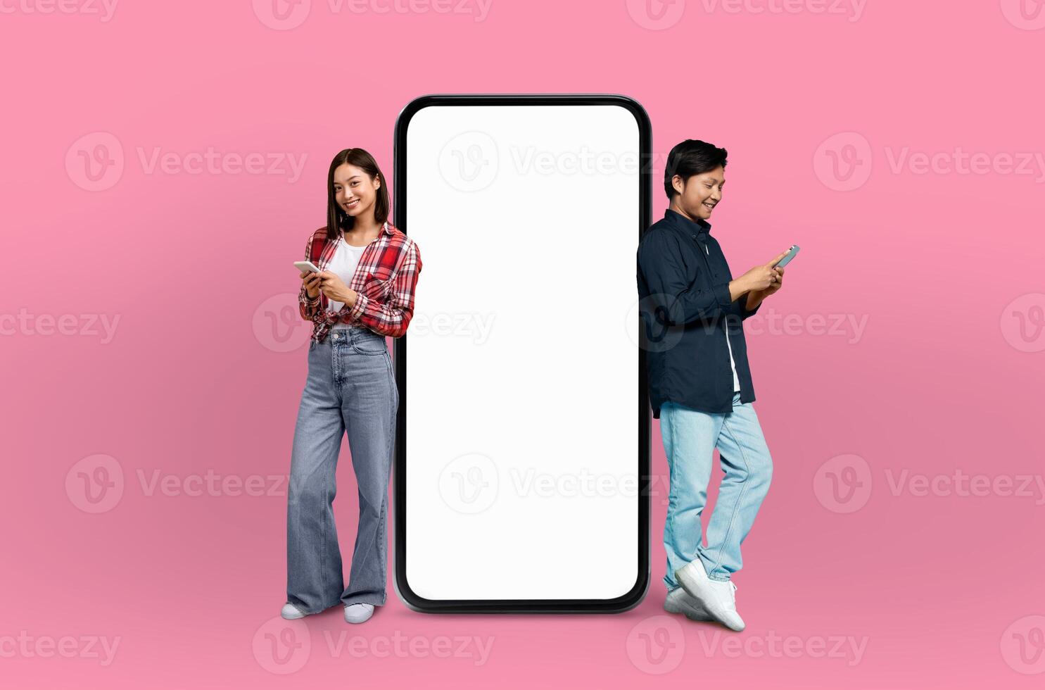 The scene features a young Chinese couple, both grinning, positioned next to a life-sized smartphone mockup on a seamless pink background. photo