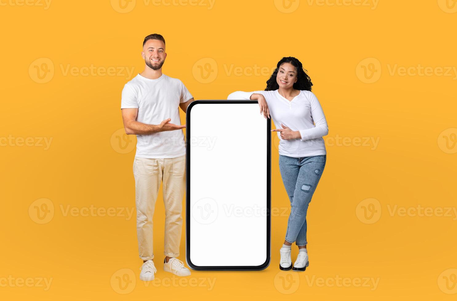 A man and a woman are standing together, holding a blank phone in their hands. They are posing against a bright yellow background, looking directly at the camera. photo