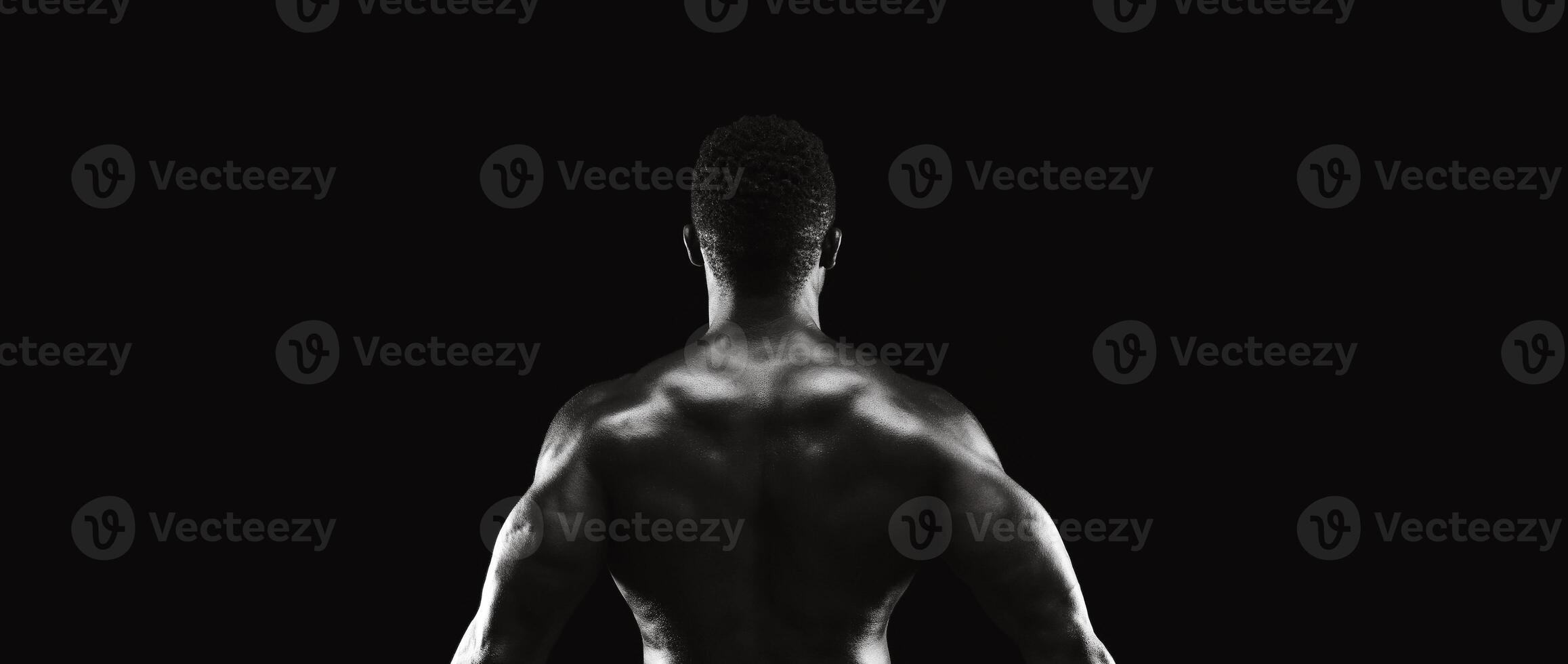 A muscular man stands with his back to the camera in a dark studio, lit by a single light source. photo