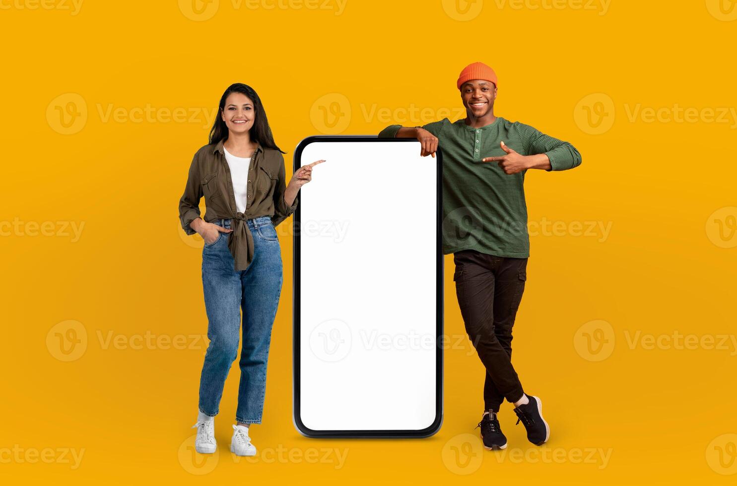 A cheerful multiracial couple stand next to an oversized, blank smartphone screen, gesturing towards it with enthusiasm, inviting attention to the copy space for advertising photo