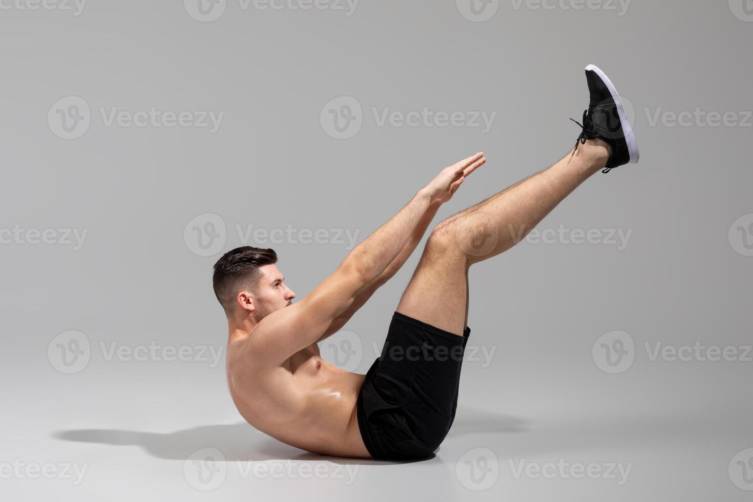 A shirtless man demonstrates the hollow body hold exercise while wearing black shorts and sneakers. The studio has a plain gray backdrop, emphasizing the form and position of the exercise. photo