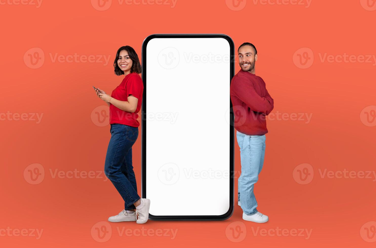 Multiethnic couple stand on either side of a giant, blank smartphone screen, both smiling and engaging with the empty display photo