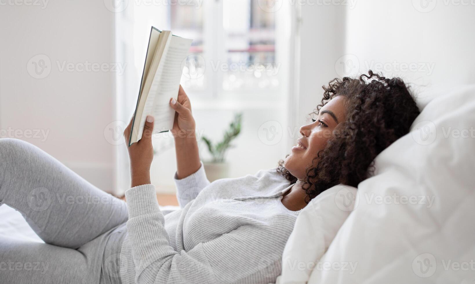 A Hispanic woman is lying down in bed with a book in hand, engrossed in reading. The room is dimly lit, and she appears relaxed and focused on the pages of the book, side view photo