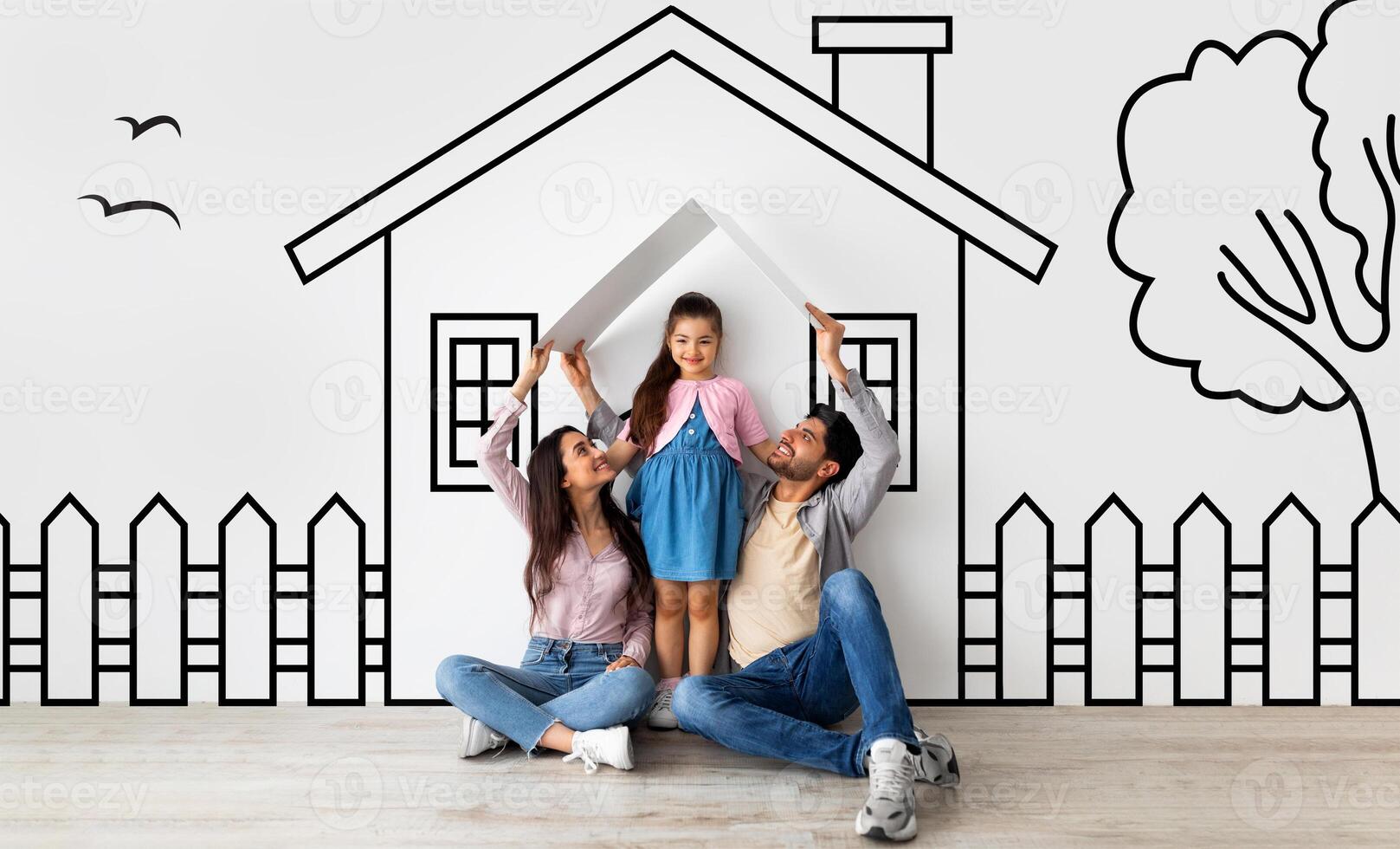 Happy middle eastern family of three father mother and daughter sitting on floor over illustrated house of their dreams over white wall background, parents holding a roof photo