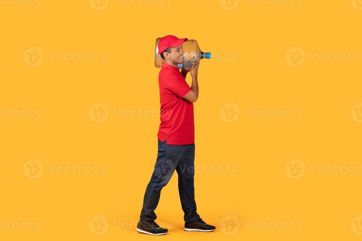 Drlivery man in a red shirt and cap carrying a large water gallon over his shoulder while standing against a solid bright yellow background. photo