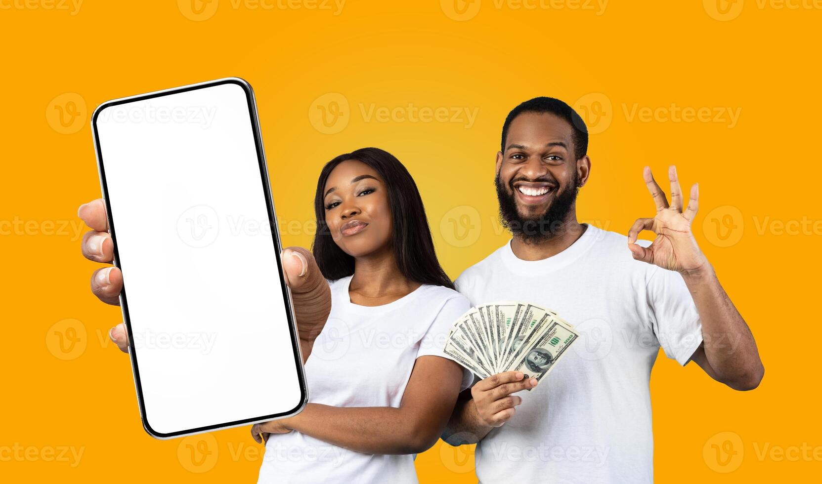 A happy black couple poses against a vibrant yellow backdrop. The woman showing smartphone in her hand, displaying a blank screen, while the man holds a fan of cash and makes the okay gesture photo