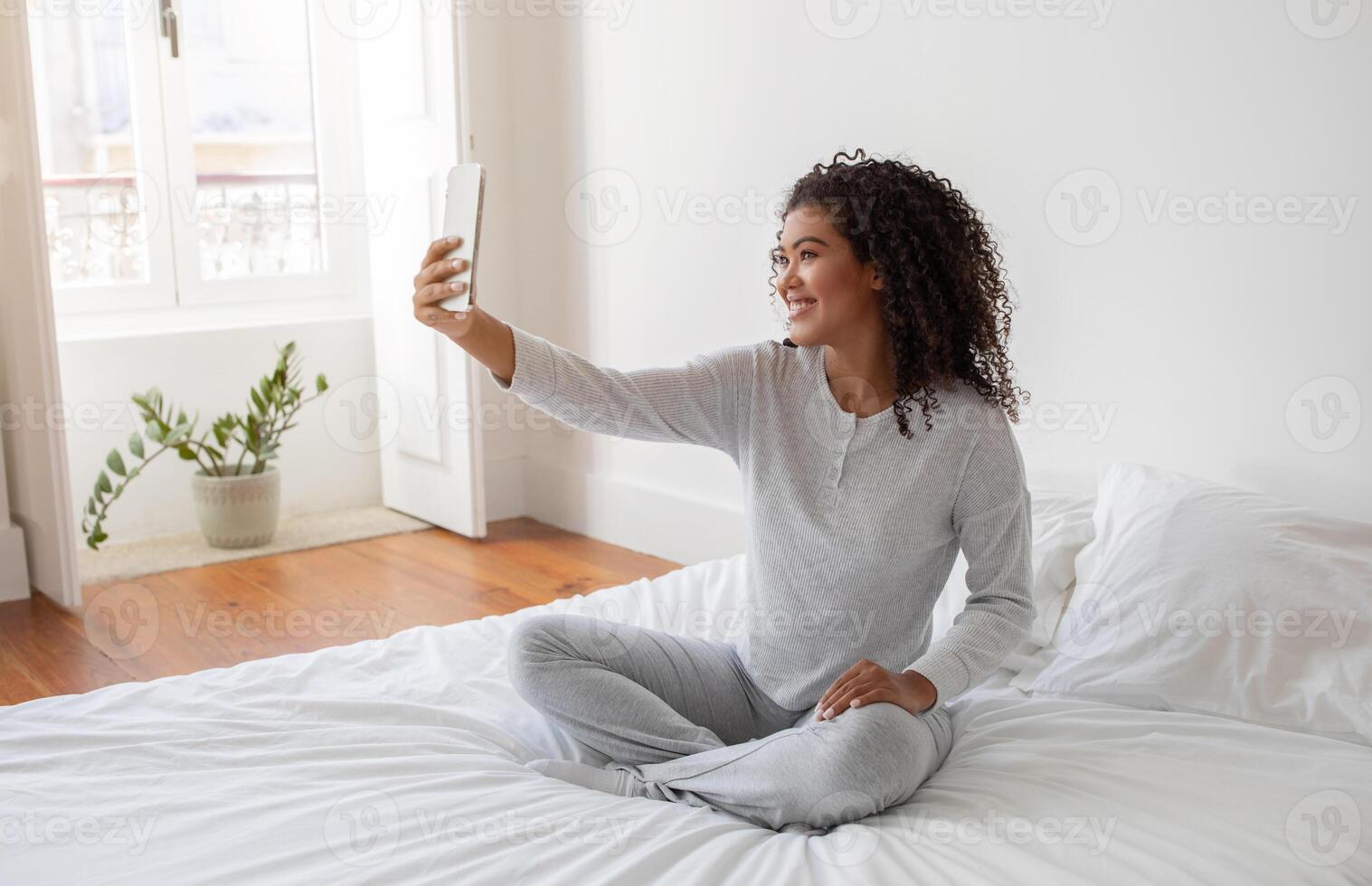 Hispanic woman seated on top of a bed, holding a smartphone at arms length to capture a selfie. She is focused on the screen, ensuring the perfect shot while sitting comfortably on the bed. photo