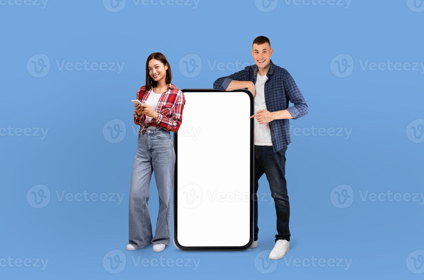 Multiethnic man and woman are standing next to a blank screen. The man is wearing a blue shirt and the woman a black dress. Both are looking at the screen with serious expressions. photo