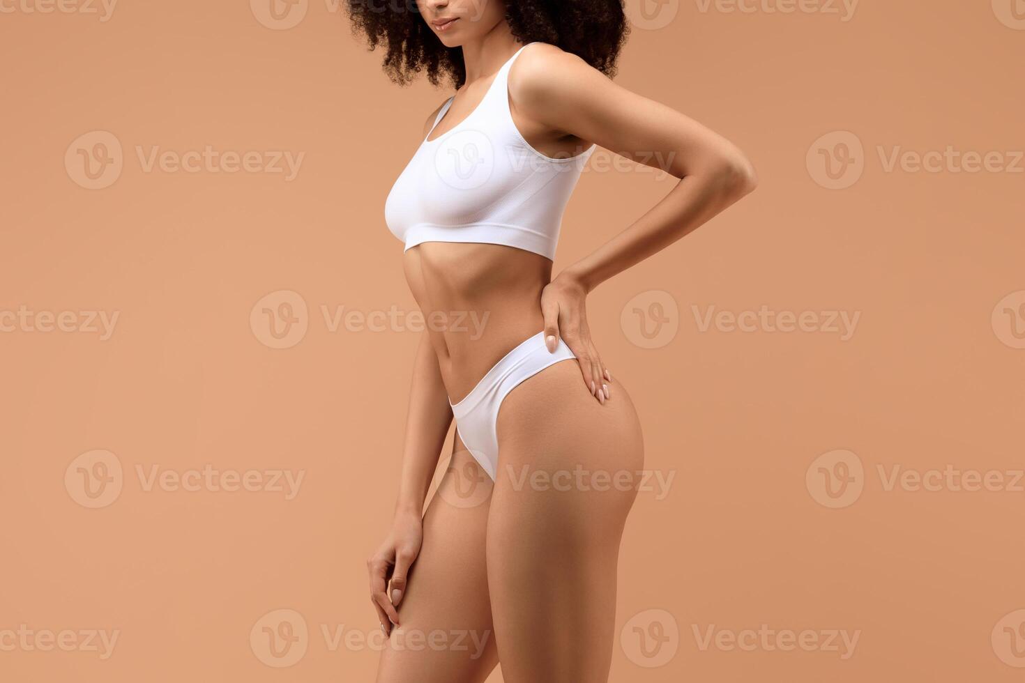 Cropped of woman with curly hair poses in white underwear against a tan backdrop, with her left hand on her hip and her right hand resting on her thigh. photo