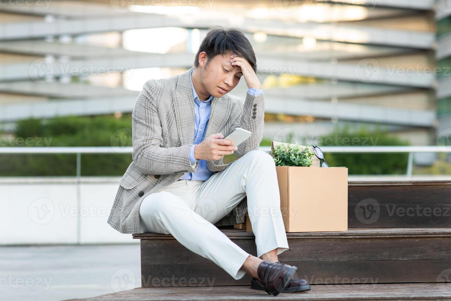 Asian young man in a blazer and white pants sits on a set of stairs, holding his phone and looking down at it with a worried expression. He has a cardboard box beside him. photo