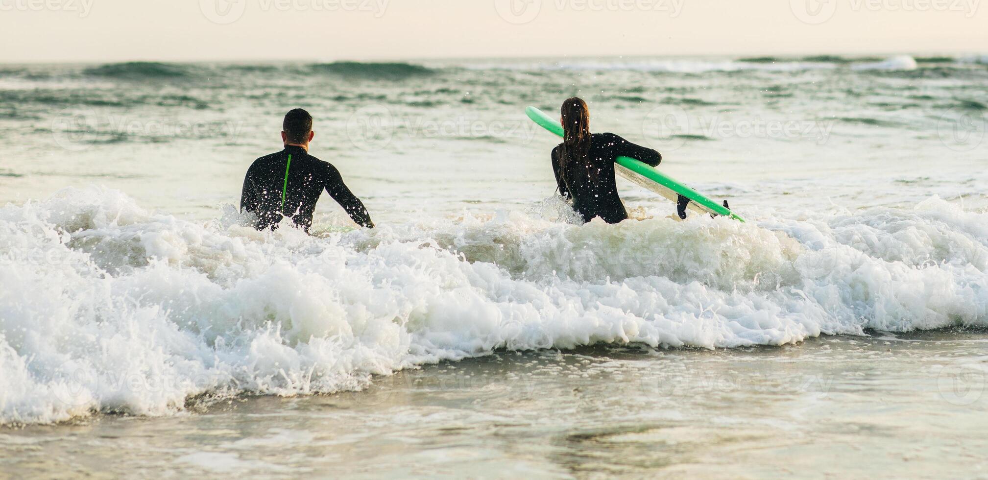 This image shows two surfers entering the ocean at sunset. They are wearing wetsuits and carrying surfboards. The waves are breaking in the background and the sky is beautiful photo