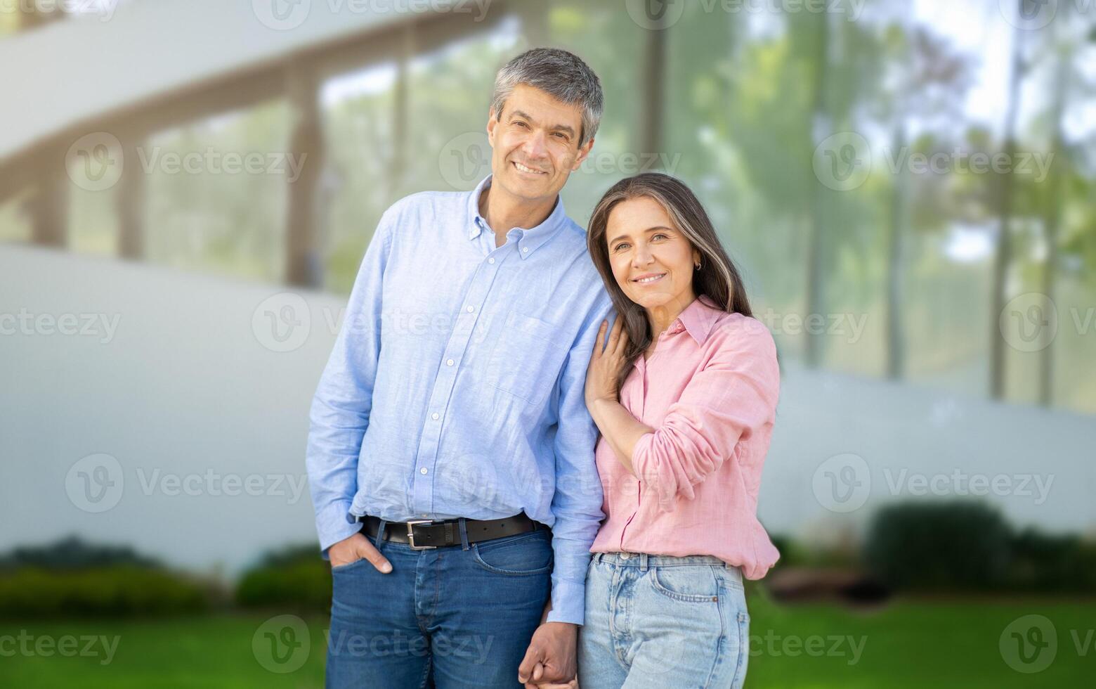 A joyful couple stands close together, sharing smiles in front of a striking modern building. The sunlight creates a warm atmosphere, highlighting their connection and happiness. photo
