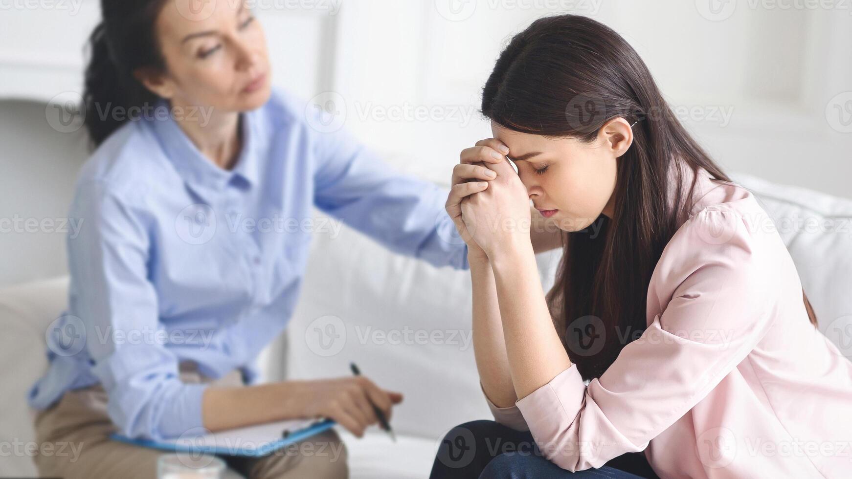 A young woman sits with her head in her hands, visibly distressed, while a therapist observes and offers comfort in a serene, private counseling room. photo