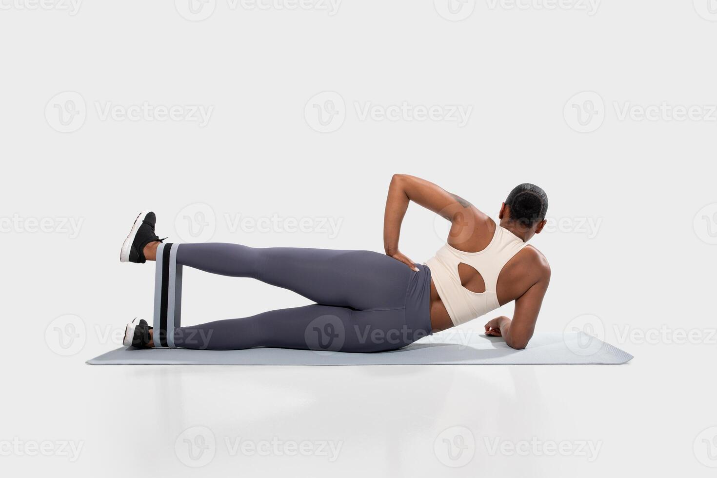 African American woman is lying on a mat, facing away from the camera, and performing a leg raise exercise with a resistance band on her thigh. She is wearing grey leggings and a white tank top. photo