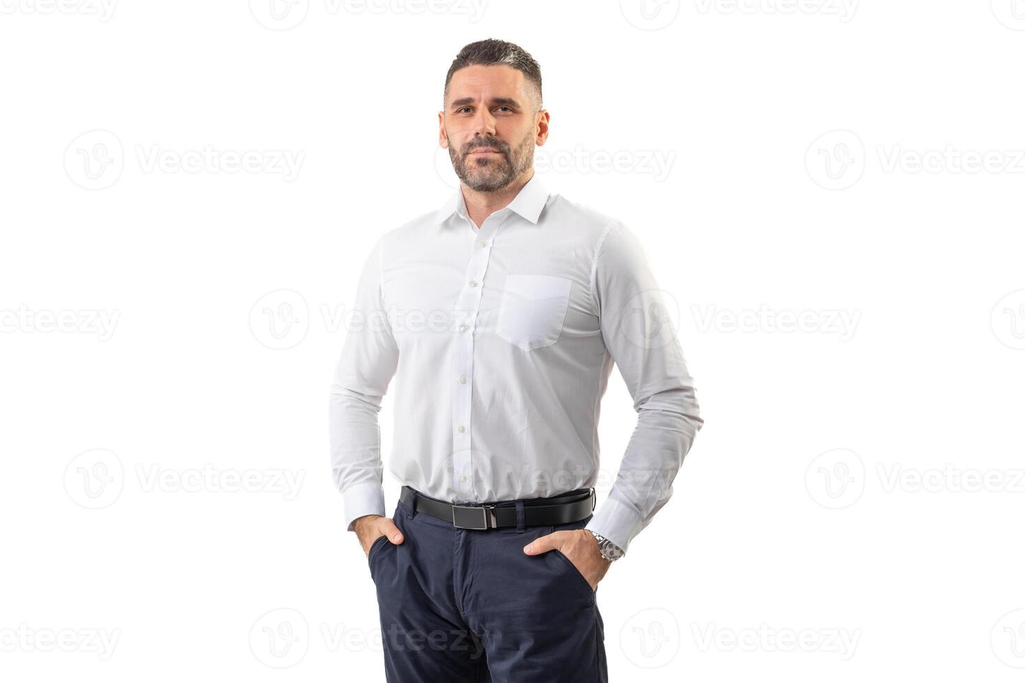 A man dressed in a crisp white shirt and dark trousers stands confidently with hands in pockets, looking directly ahead. He is captured against a plain white backdrop photo