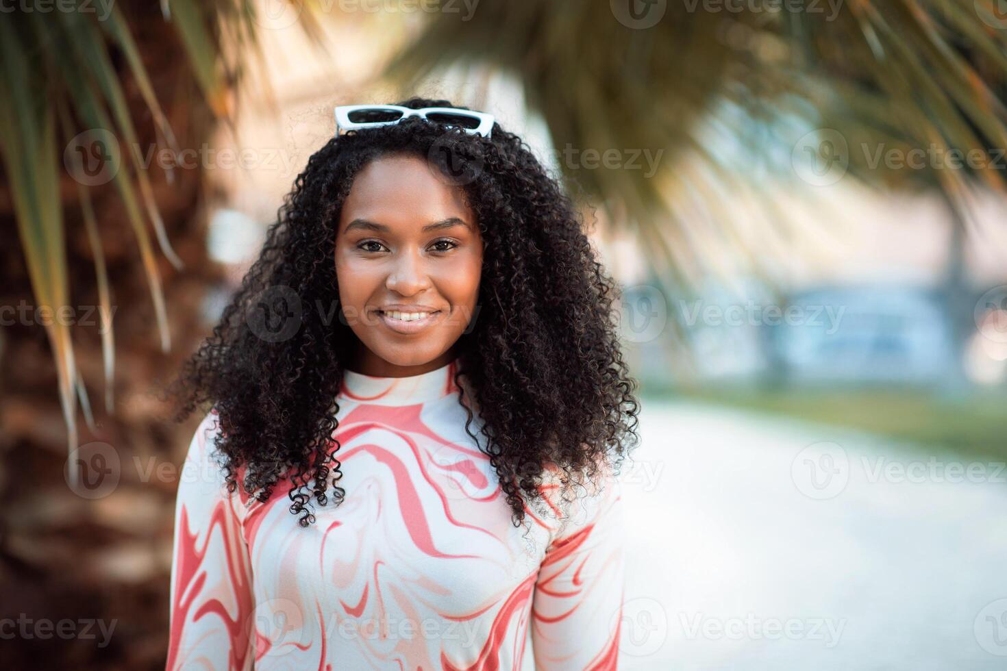 This image shows a woman with long, curly black hair smiling in front of palm trees. She is wearing a white and pink shirt with a marbled pattern and has a pair of sunglasses on top of her head. photo