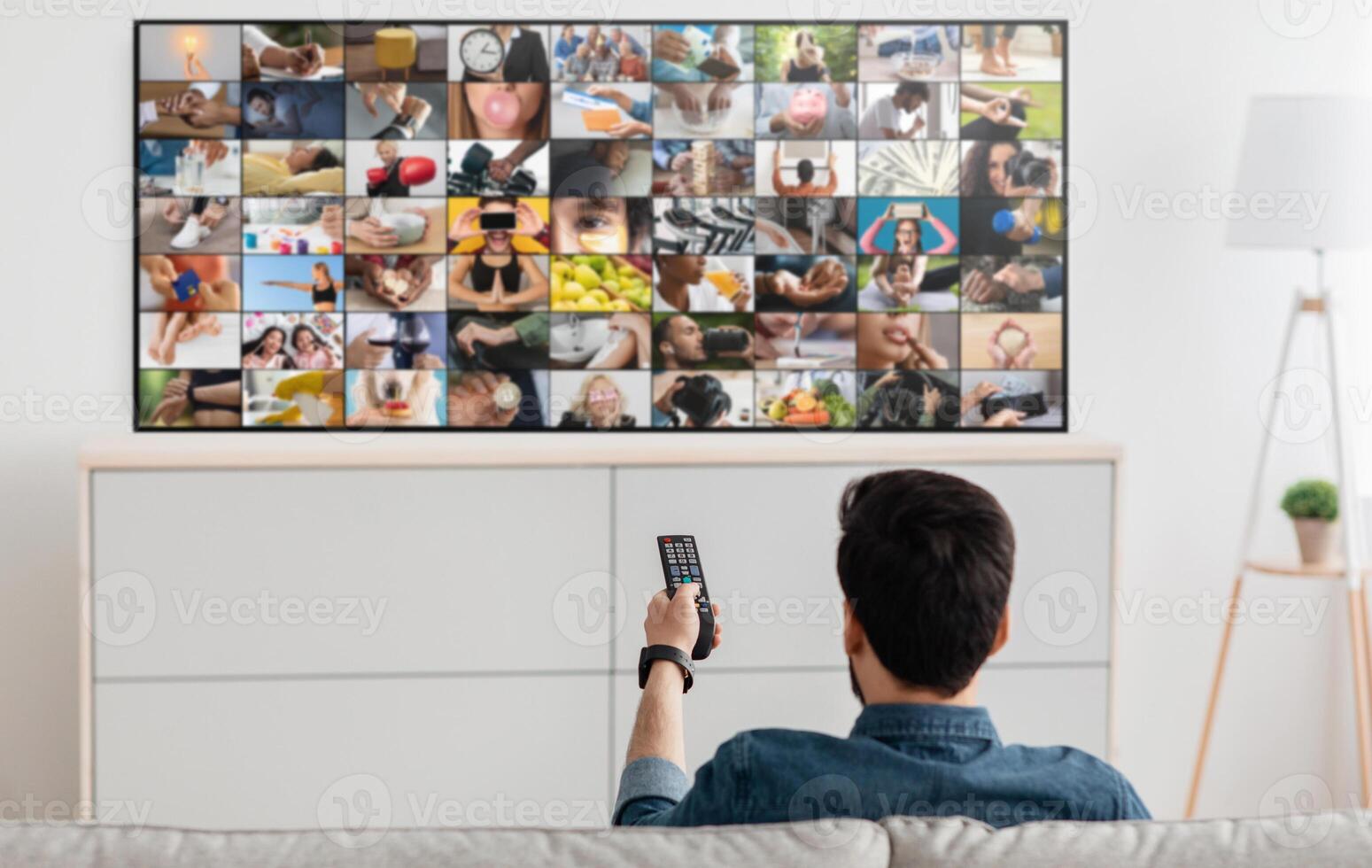 A man sits on a couch and watches television, holding the remote in his right hand. The TV screen displays a collage of various images, including people, objects, and scenes photo