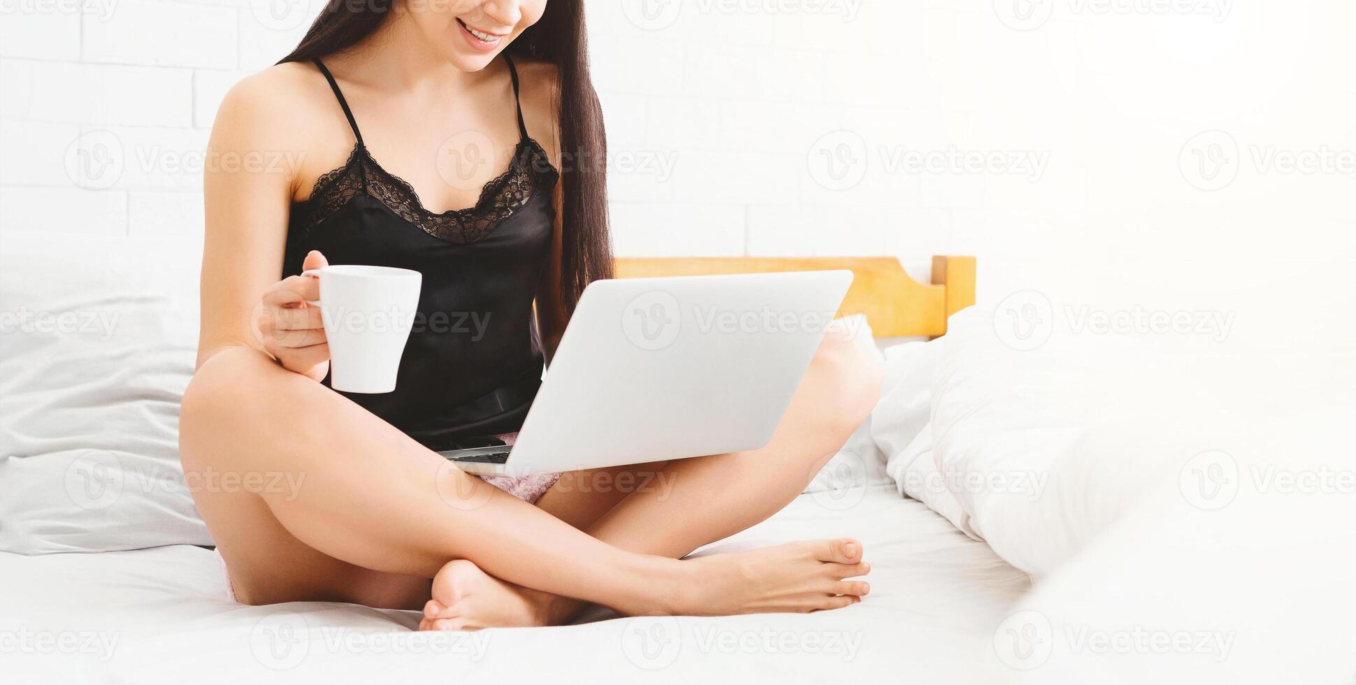 A woman is seated on a bed, holding a cup in one hand and a laptop in the other. She appears focused on the screen, possibly working or browsing. The room is simple, with neutral decor. photo