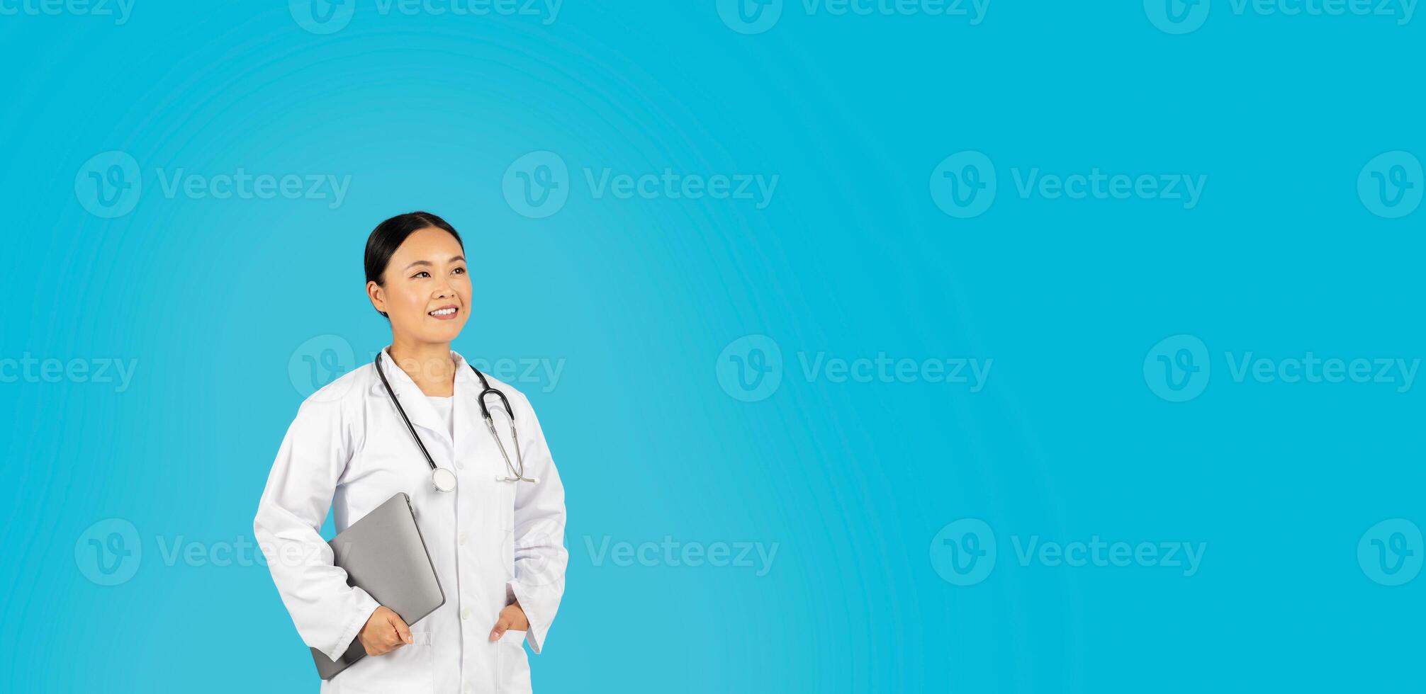 Asian woman doctor, wearing a white coat and a stethoscope, smiles while holding a laptop in her left hand and looking at copy space. She is standing in front of a light blue background. photo