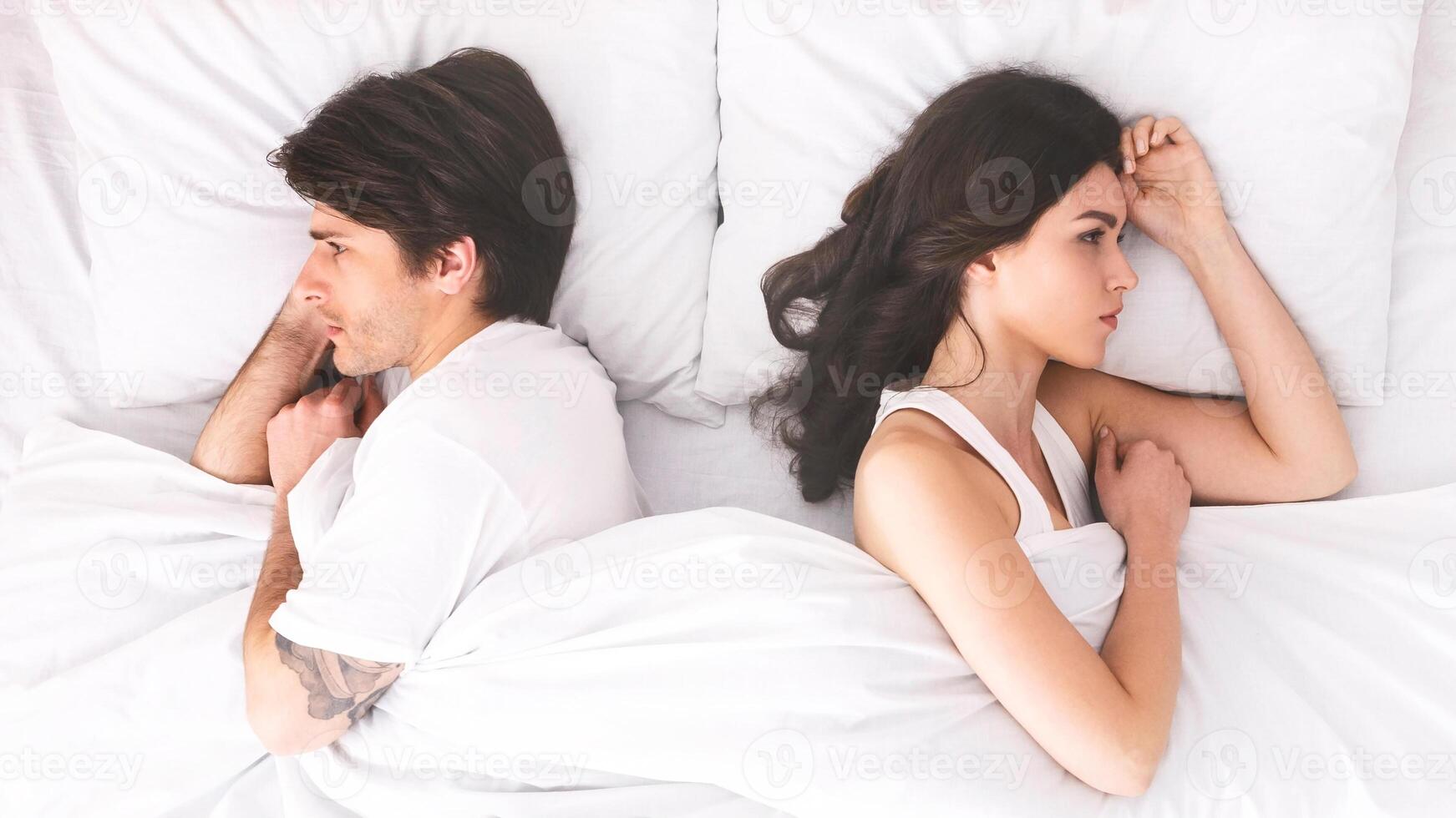 A young couple lies in bed, turned away from each other, seemingly after an argument. They are both lying on their sides, with their backs to each other photo