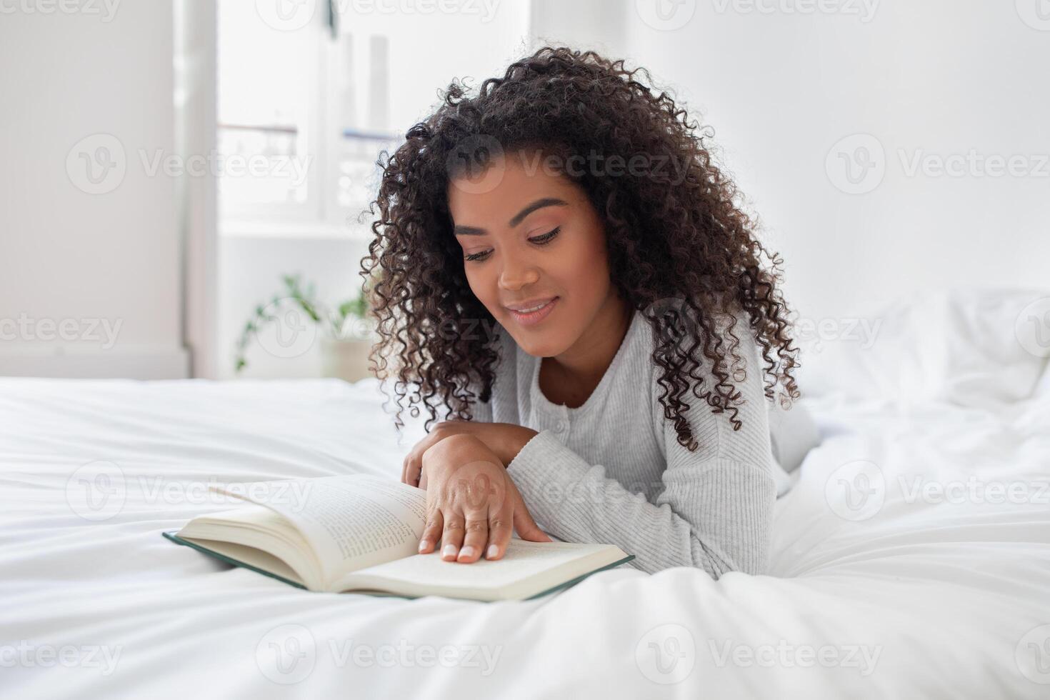 A Hispanic woman is laying comfortably on a bed, engrossed in reading a book. The room is softly lit, and she is fully focused on the text, turning pages occasionally. photo