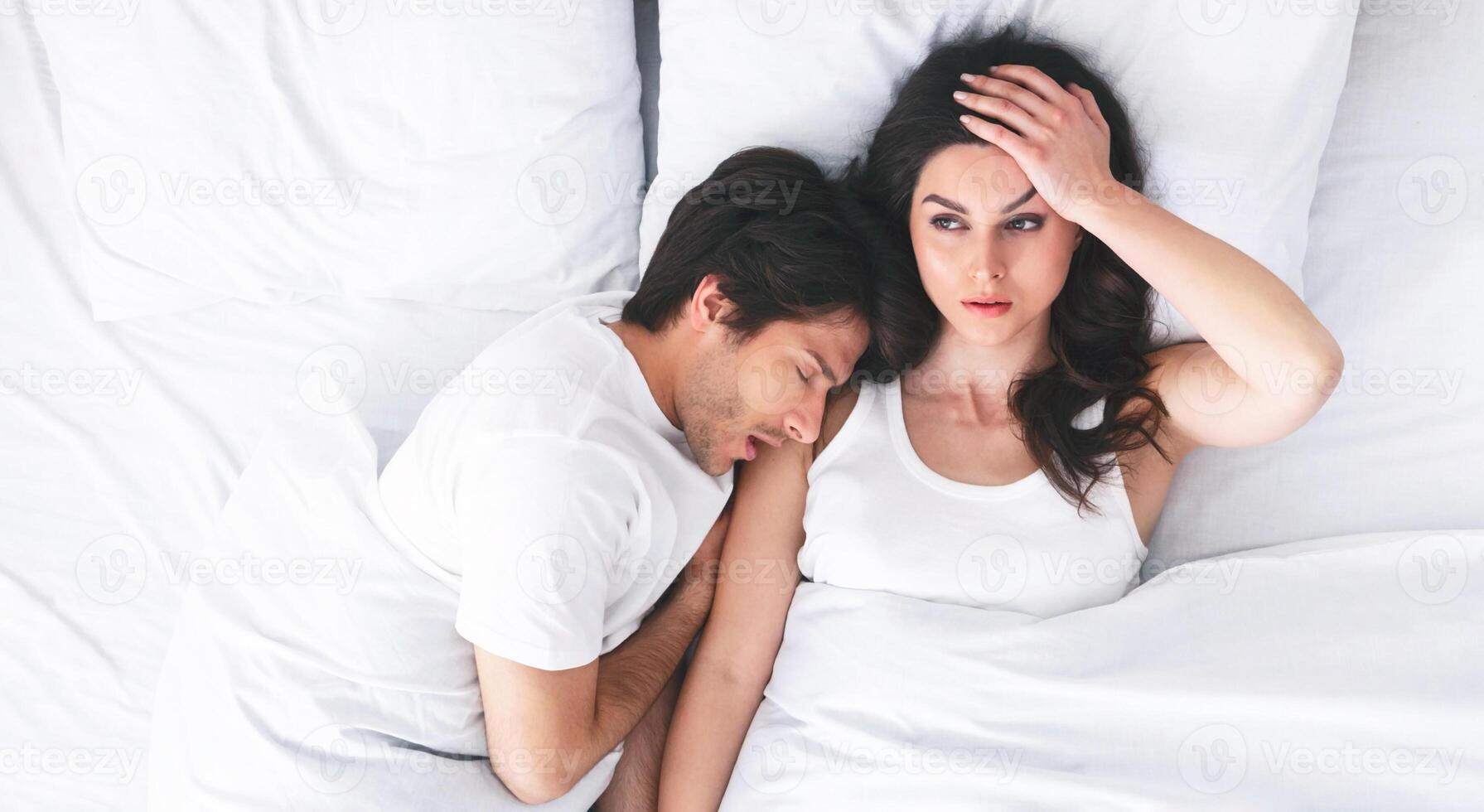 A young woman lies awake in bed with a frustrated expression, while her partner sleeps soundly beside her. Her hand is on her forehead, as if she is trying to block out the noise. photo