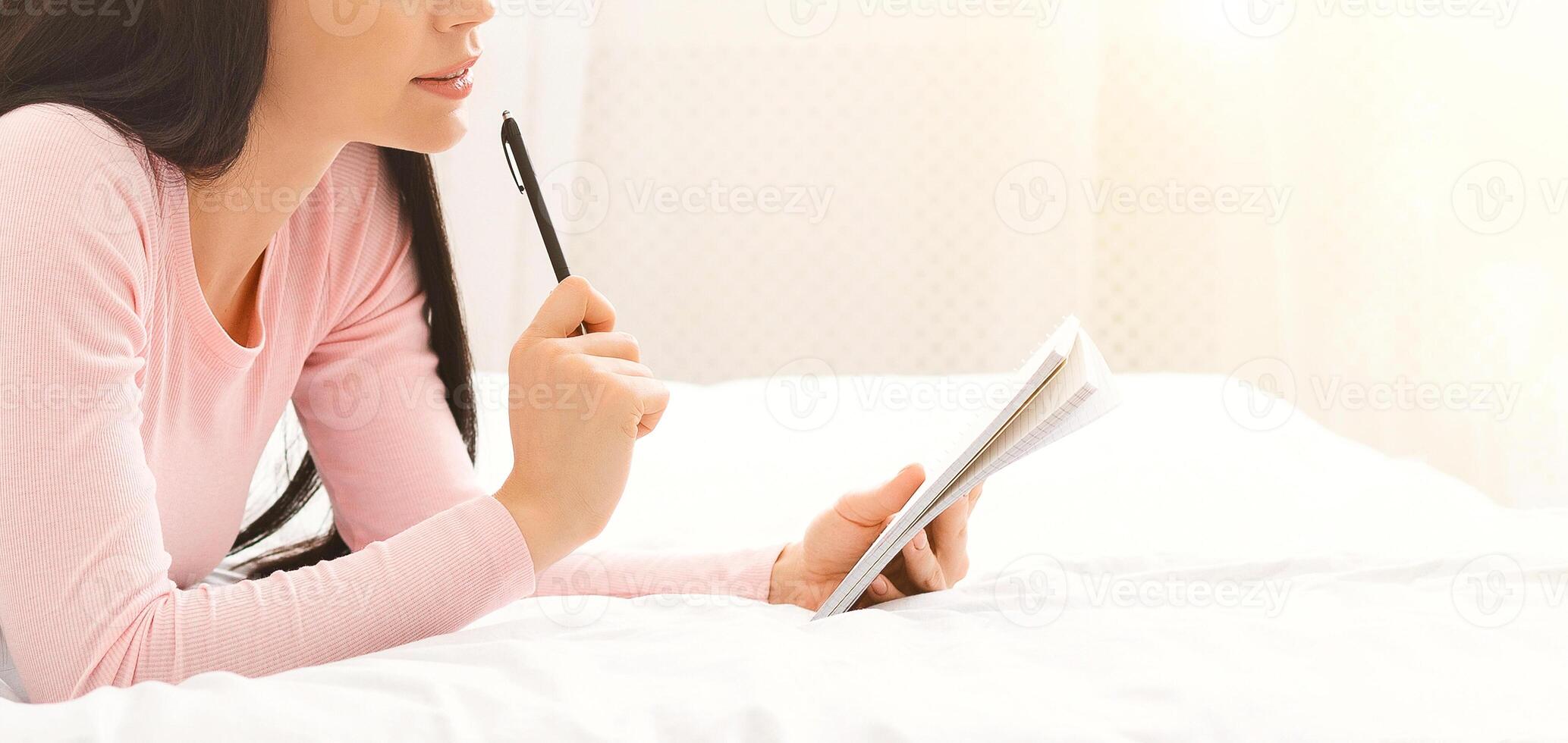 A young woman is seated indoors with natural light illuminating the room. She appears focused and thoughtful as she writes in a notebook with a pen, cropped, panorama with copy space photo