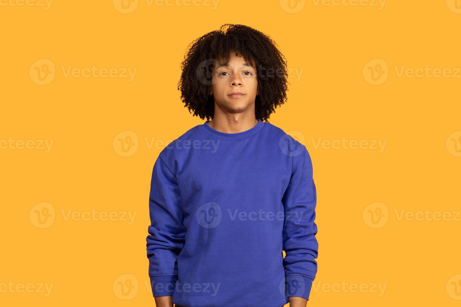 A young man with an curly hairstyle stands in front of a bright yellow background, wearing a blue sweatshirt. He looks directly at the camera with a neutral expression. photo