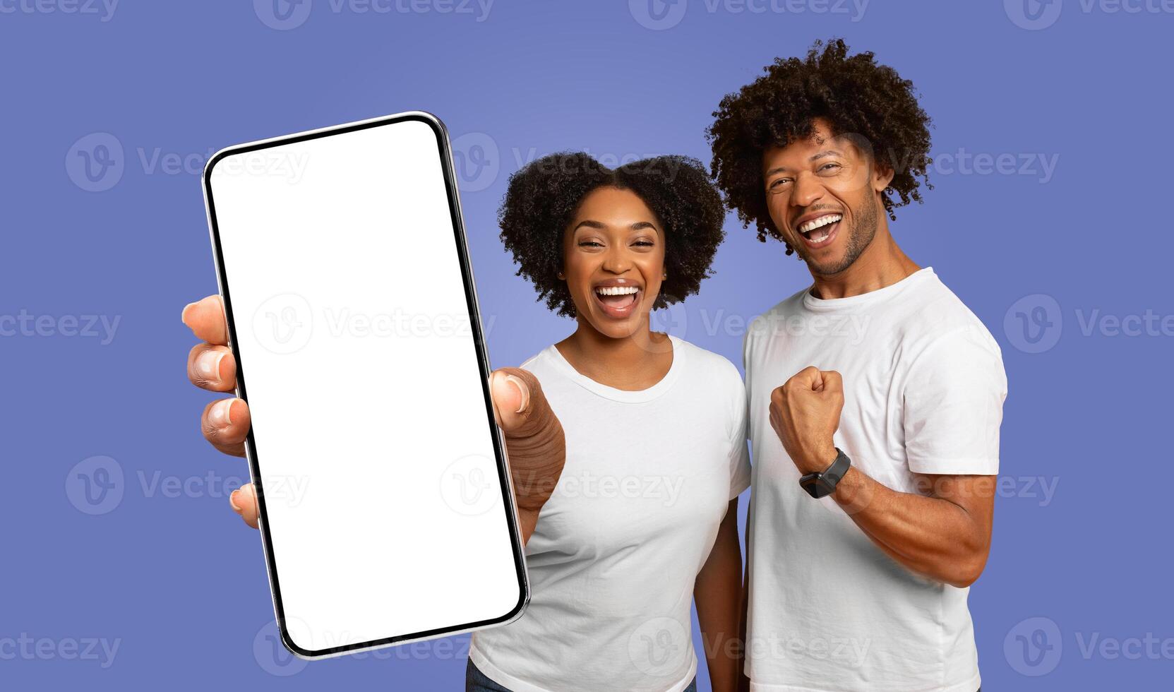 A happy African American couple stands in front of a purple background, with the woman holding a smartphone with a blank screen towards the camera and the man raising a fist in celebration photo