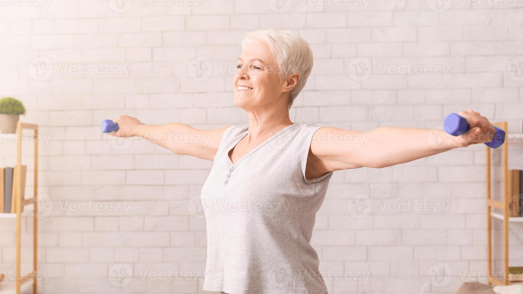 Senior woman in athletic wear is performing various exercises with dumbbells in a brightly lit room. She is focused and determined, showcasing strength and stamina during her workout routine. photo