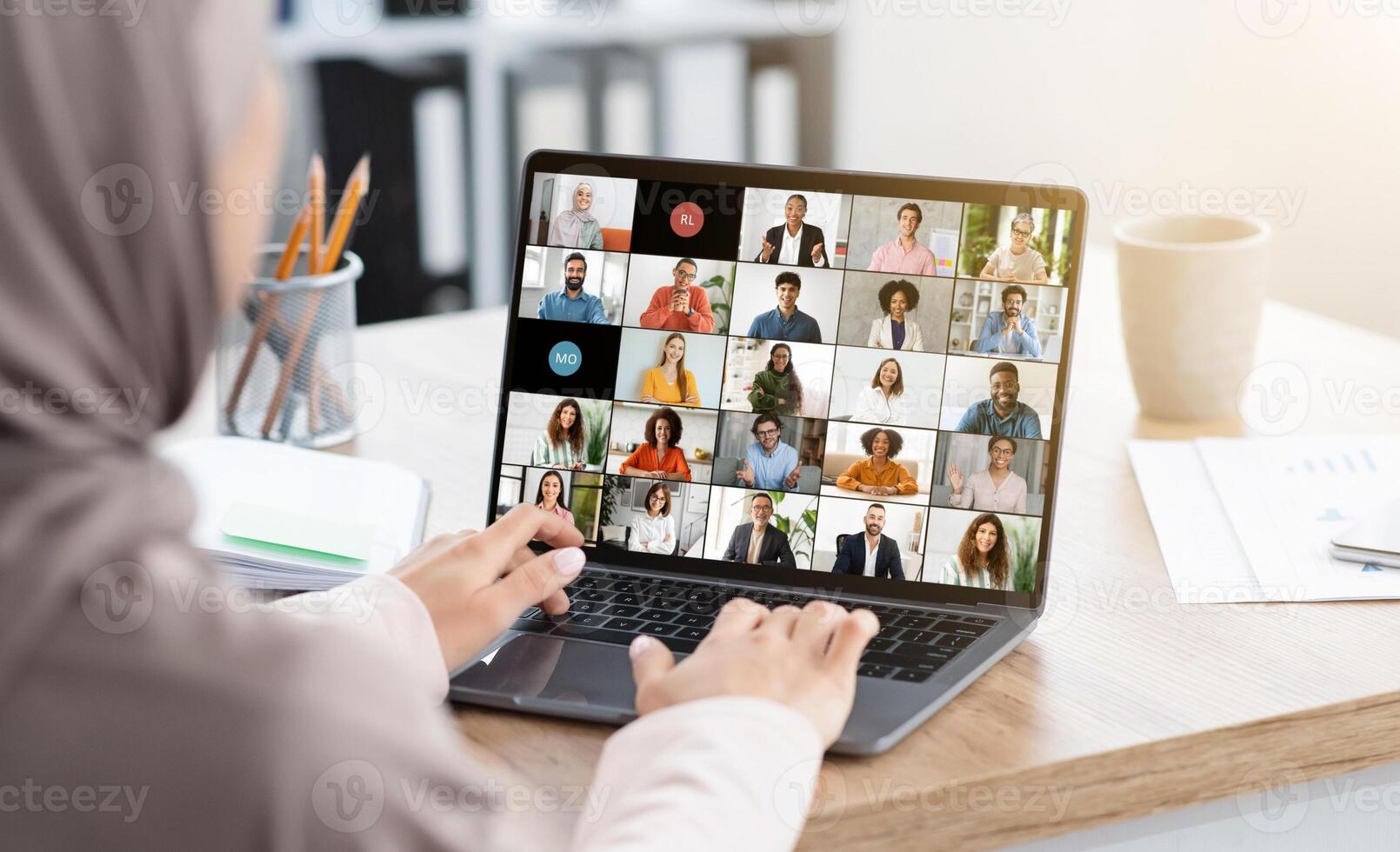 Muslim woman sits at a desk in a home office setting, participating in a large virtual meeting on her laptop. The screen is filled with tiles of diverse people participating in the meeting photo