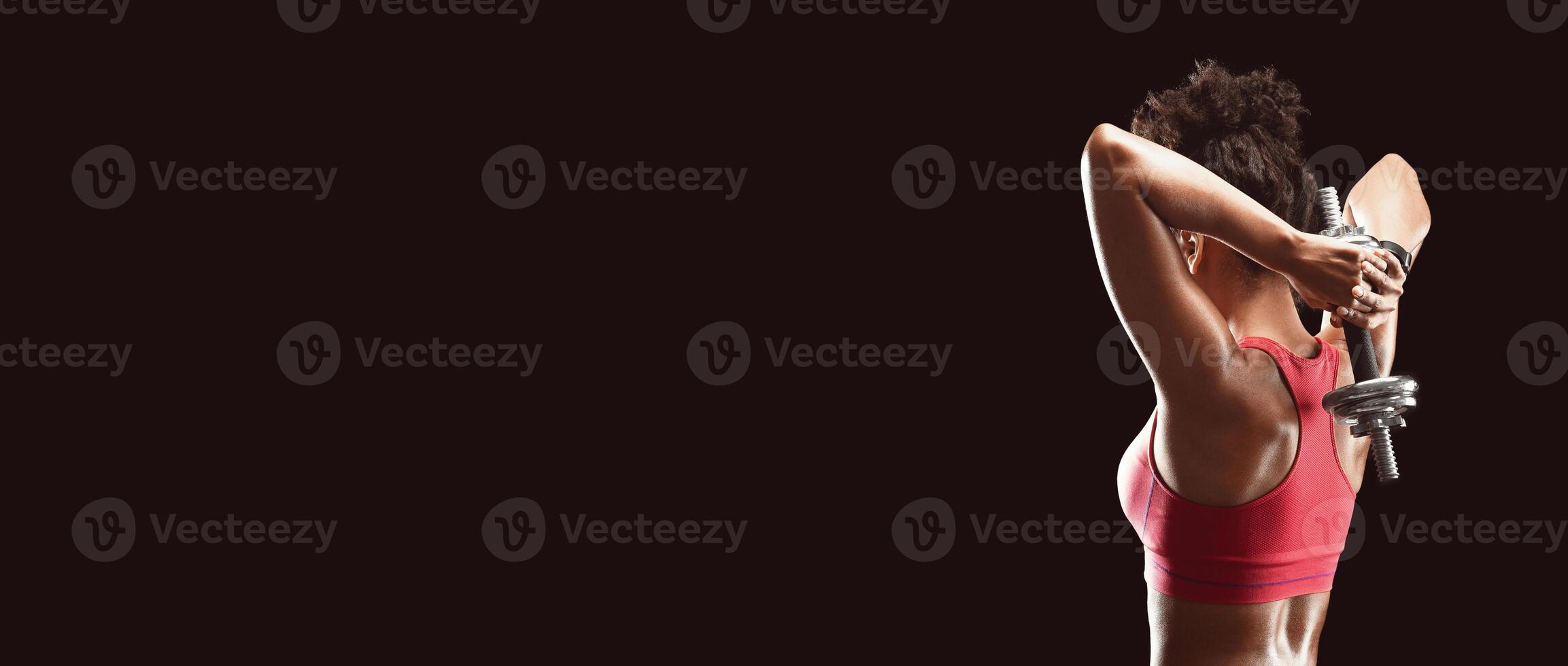 African athletic girl working on her back muscles, using barbell over black background, empty space photo