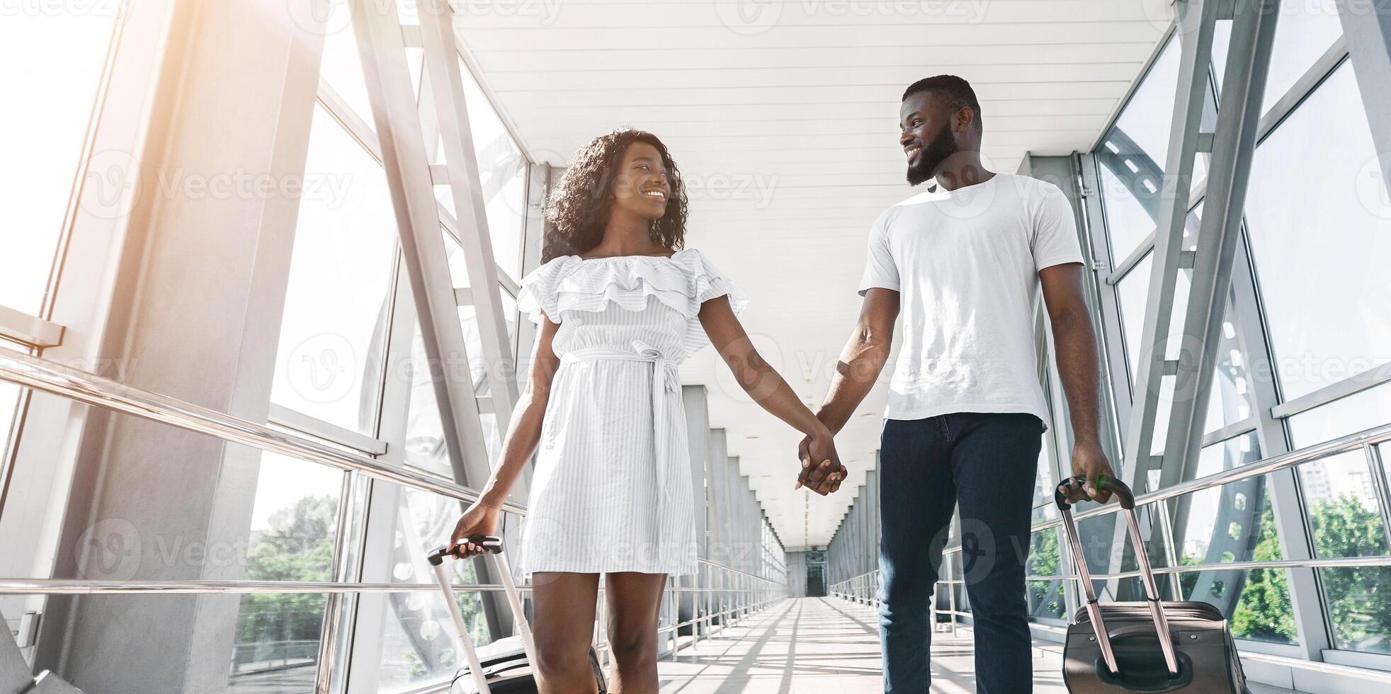 Black couple walks hand-in-hand through an airport terminal, smiling and happy. They are both pulling wheeled luggage and look excited for their upcoming vacation, copy space, sun flare photo