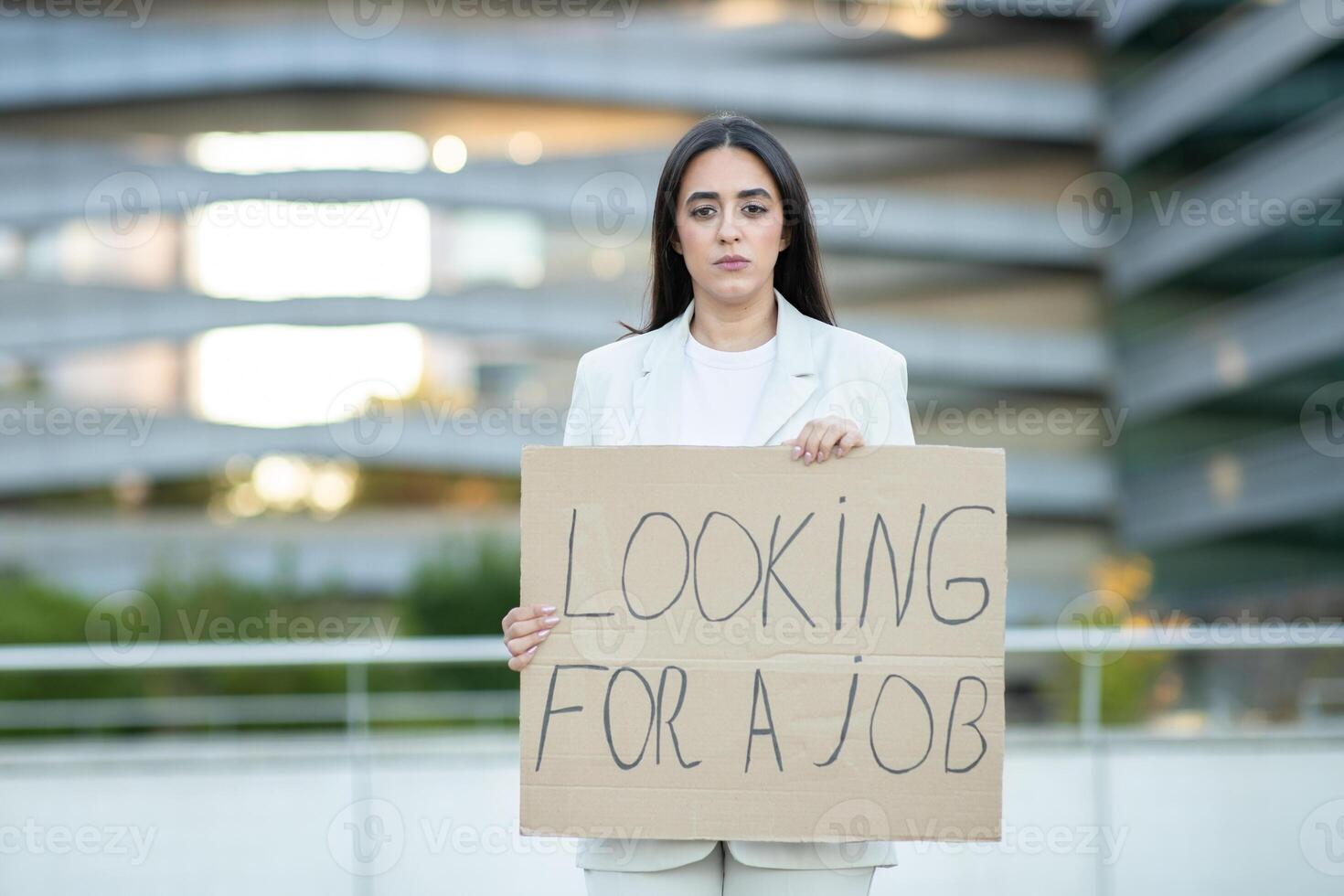 A young woman stands in an urban setting, holding a large cardboard sign that reads Looking for a job, expressing her search for employment. photo