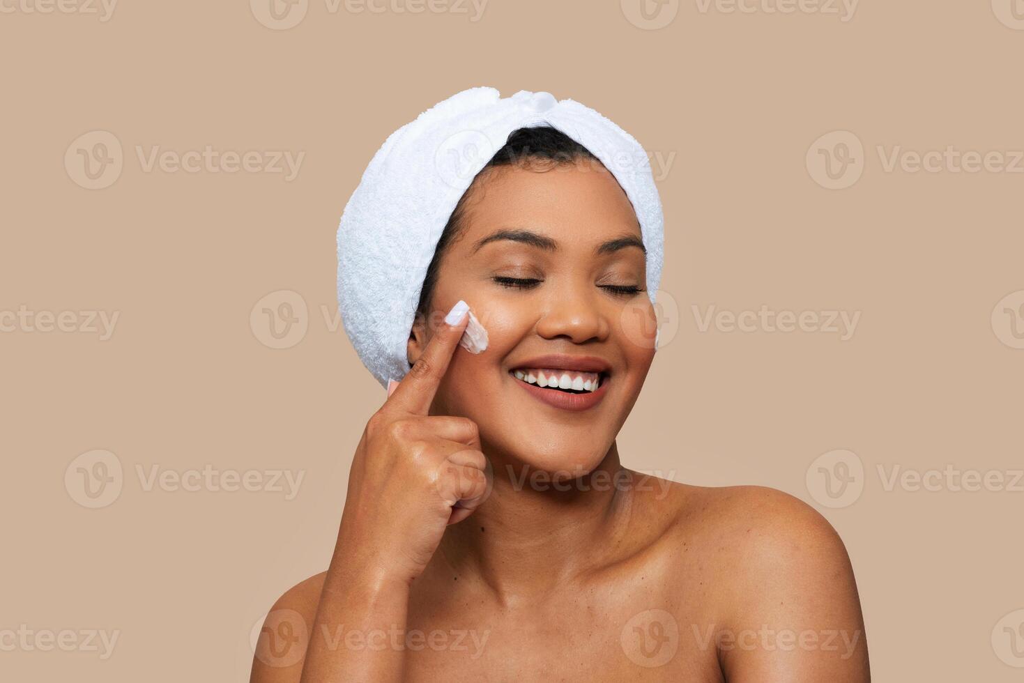 A woman with a towel wrapped around her head is gently applying cream to her face. She is standing in front of a mirror, focusing on her skincare routine. photo