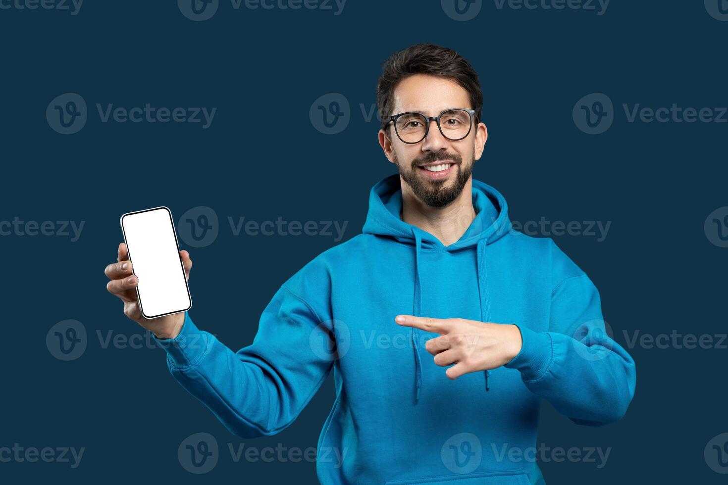 A cheerful young man with a beard and glasses is wearing a blue hoodie and presenting a smartphone with its screen facing forward towards the viewer. He is pointing at the screen photo
