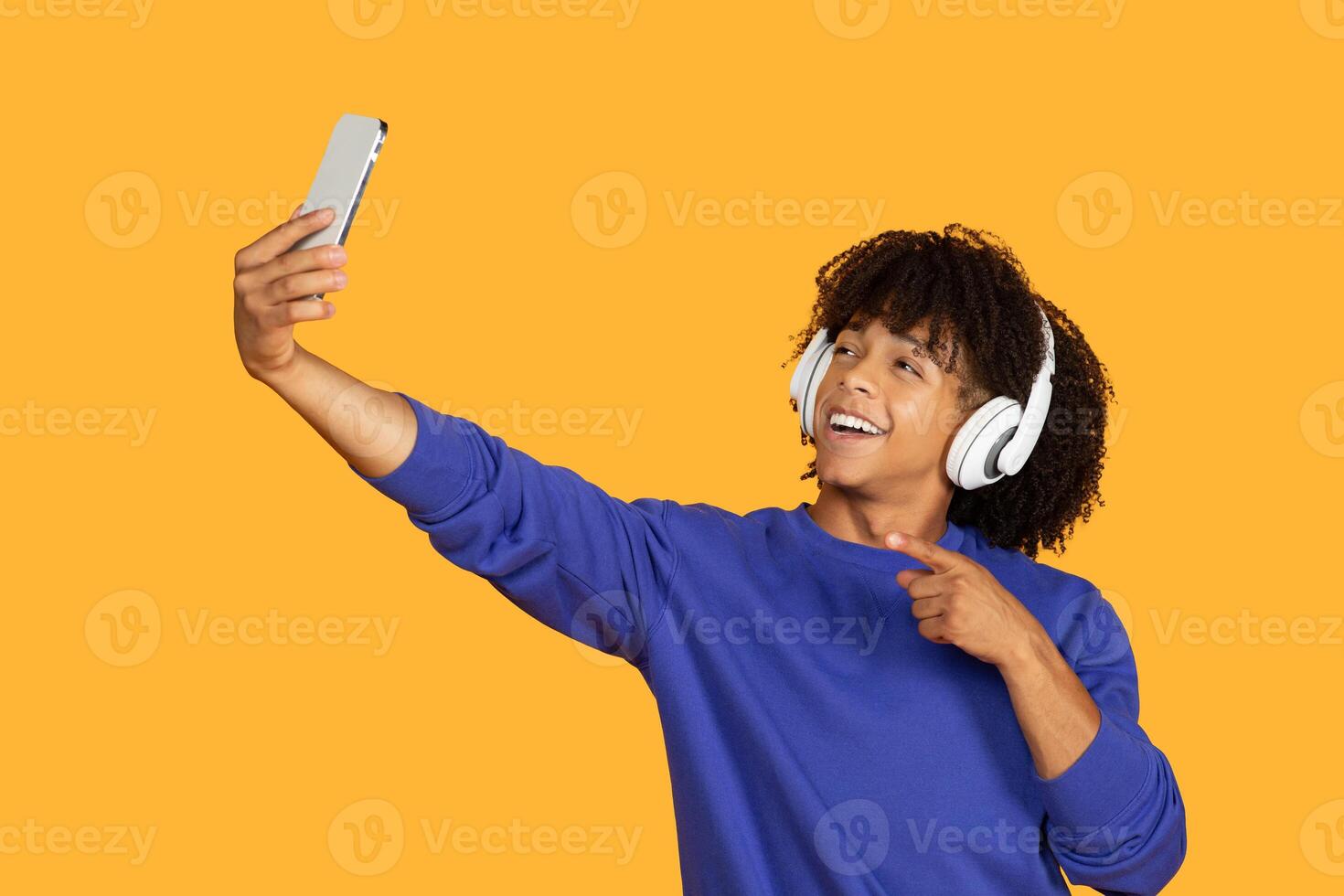 A young man with curly hair is taking a selfie on a yellow background. He is wearing headphones and smiling happily. His index finger is pointed towards the camera as if he is giving a thumbs up. photo