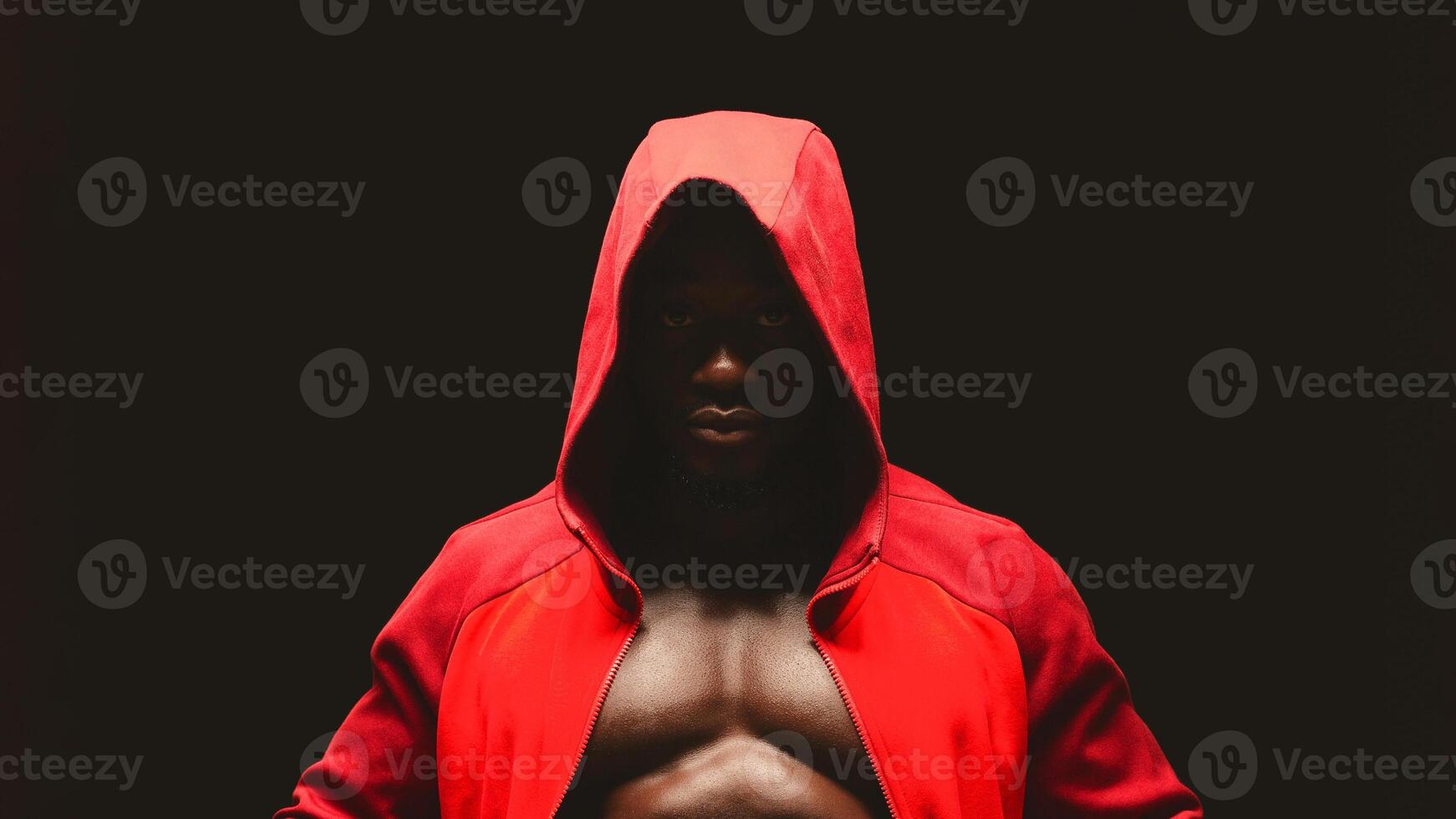 A man wearing a red hoodie stands with a serious expression against a dark background, his chest muscles visible. photo
