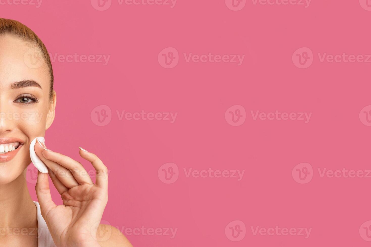 A young woman with long blonde hair is smiling and applying a cotton pad to her cheek. She is standing in front of a solid pink background. Her expression is happy and relaxed, copy space photo