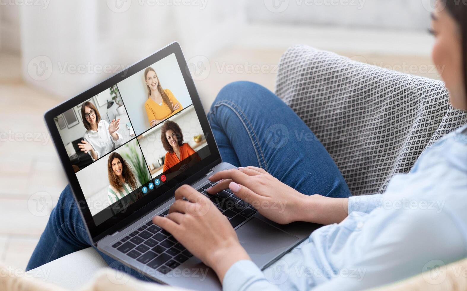 A woman is actively participating in a remote team meeting using her laptop. She is seated comfortably on a couch in a well-lit living room, engaging with four colleagues displayed on the screen. photo